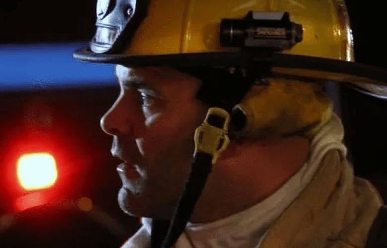 A firefighter wearing a yellow helmet and protective gear looks towards the left. Red emergency lights are blurred in the background.