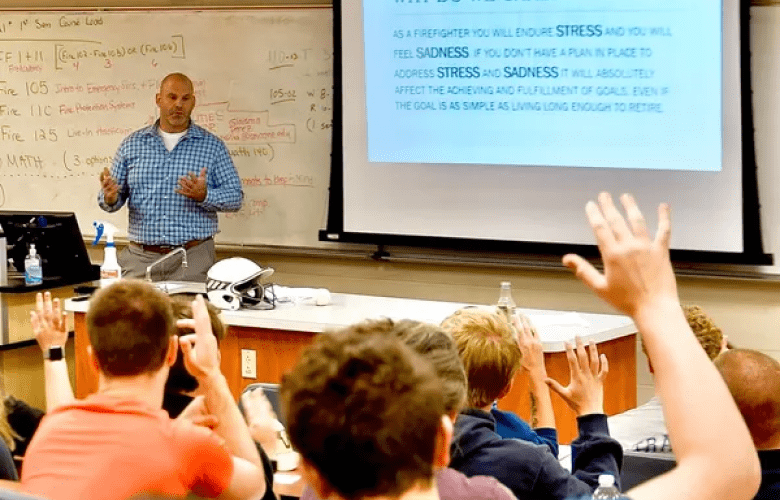 A lecturer stands at the front of a classroom next to a whiteboard and a projector screen, speaking to students who are raising their hands. The room appears to be in an educational setting.