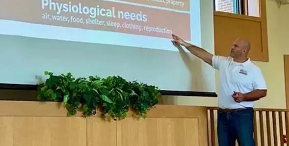 A man in a white polo shirt stands in front of a presentation screen pointing to a slide titled "Physiological needs" which lists air, water, food, shelter, sleep, clothing, and reproduction.