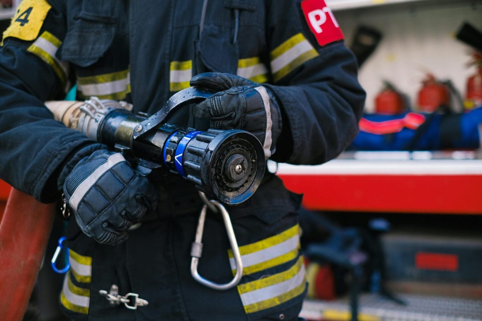 A close-up view of a firefighter holding a fire hose nozzle, dressed in protective gear with reflective stripes, near firefighting equipment.