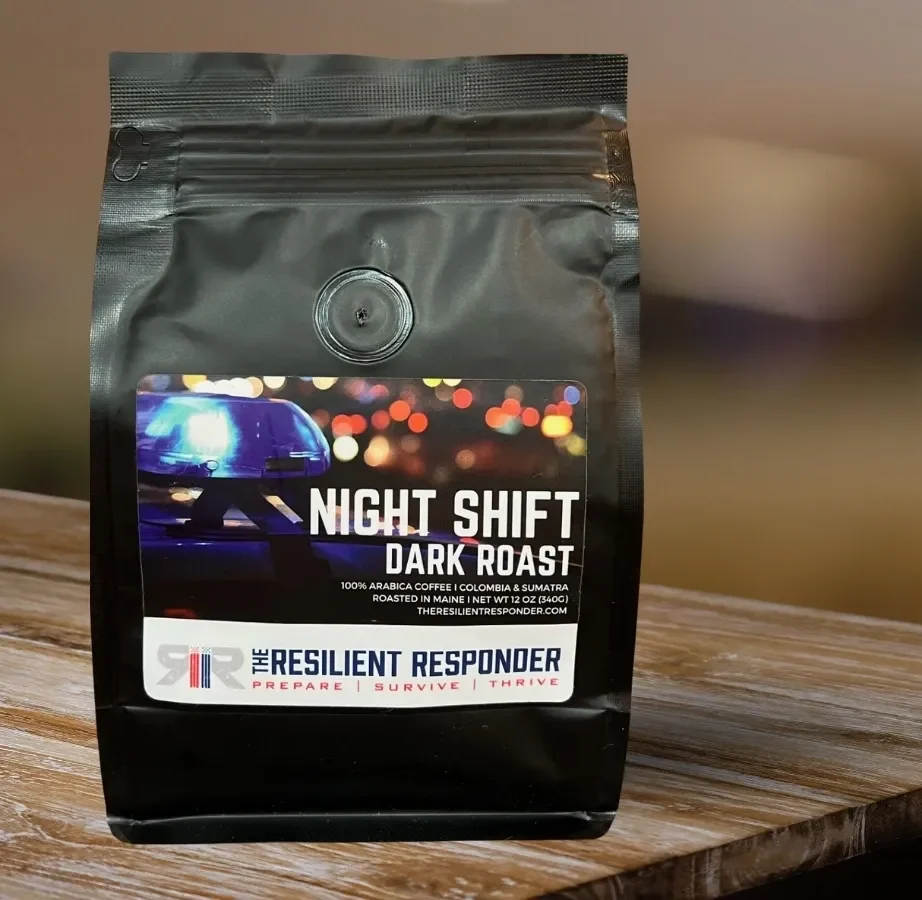 A black bag of Night Shift Dark Roast from The Resilient Responder, featuring a police car graphic. Label includes "100% Arabica Coffee" and website information. Bag on a wooden table.